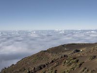 an empty road is descending above the clouds below a hill near a town on a hilltop